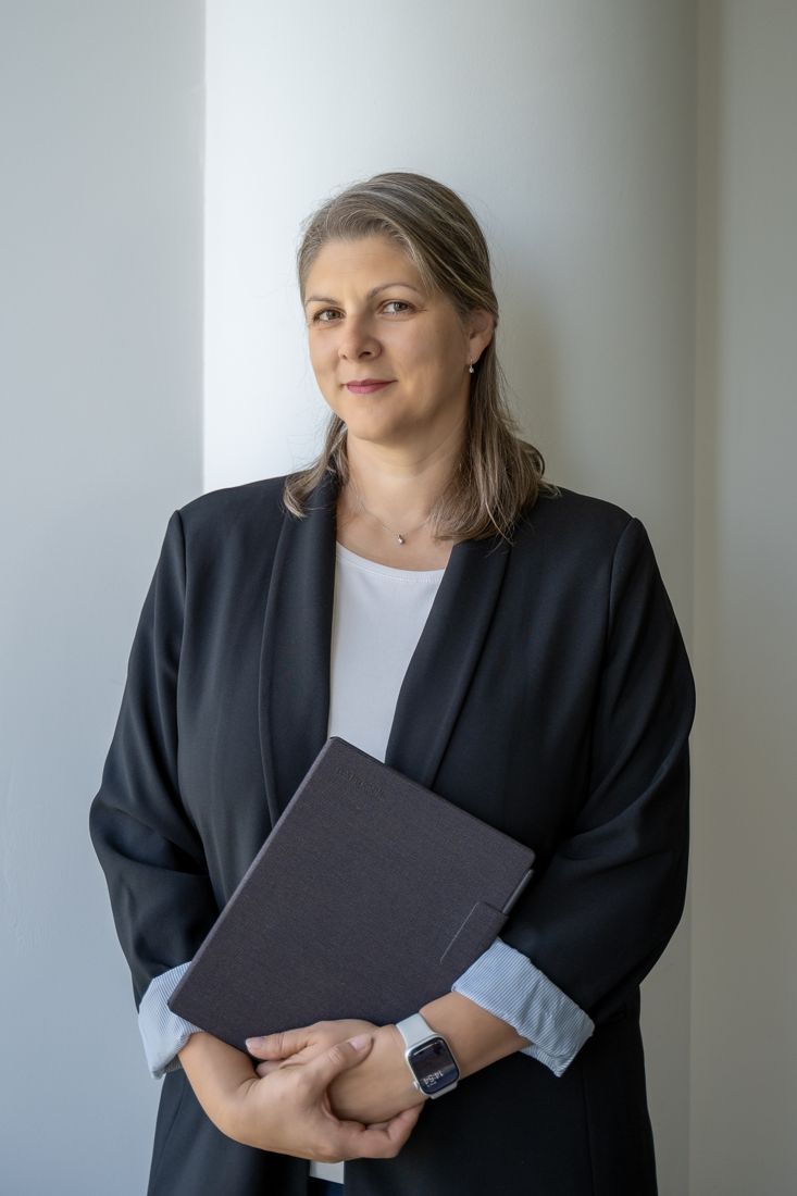 Žena v tmavom saku stojí pri okne a drží zápisník
